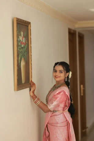 South Indian bride wearing a pink silk saree with gold jewelry touching a wall frame in an indoor hallway