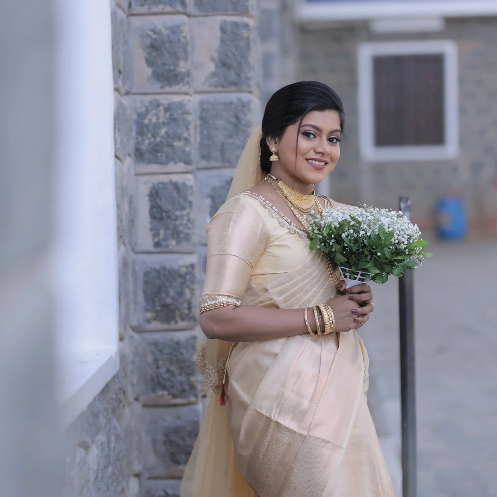 South Indian Christian bride wearing a cream gold saree, gold jewelry, and holding a bridal bouquet near a stone wall