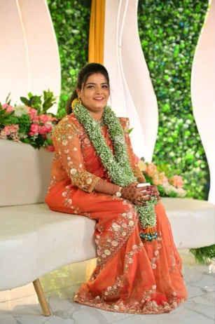 South Indian bride in an orange silk saree seated on a couch with white and green floral stage backdrop