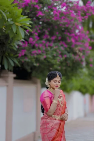 South Indian bride wearing a pink silk saree with gold jewelry and white flowers in her hair standing outdoors near magenta flowers